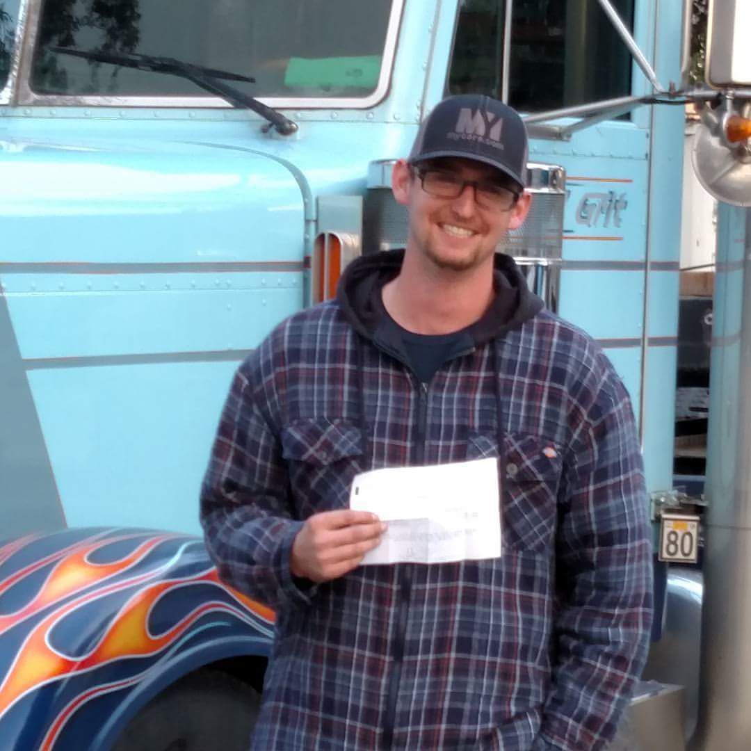 Man holding paper, standing in front of a blue semi-truck with flame decals, wearing a plaid shirt and a NY hat.