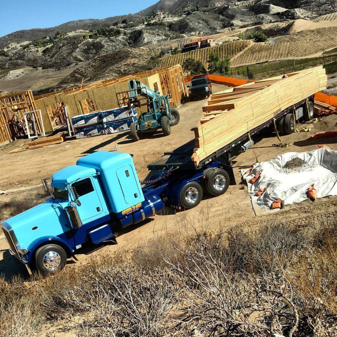 Blue semi-truck unloading lumber at a construction site on a hillside with a backhoe and house frame.