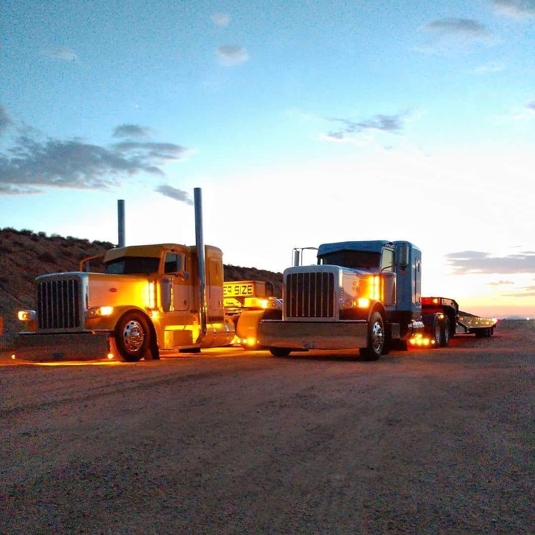 Two semi-trucks, one yellow, one blue, parked side-by-side on a dirt road at dusk.