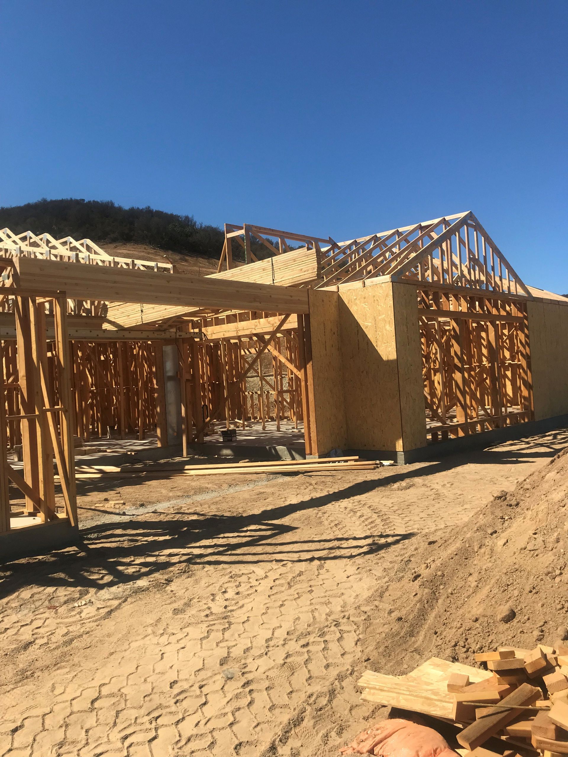 Construction site: wooden frames of a building under a blue sky, dirt foreground, hillside backdrop.
