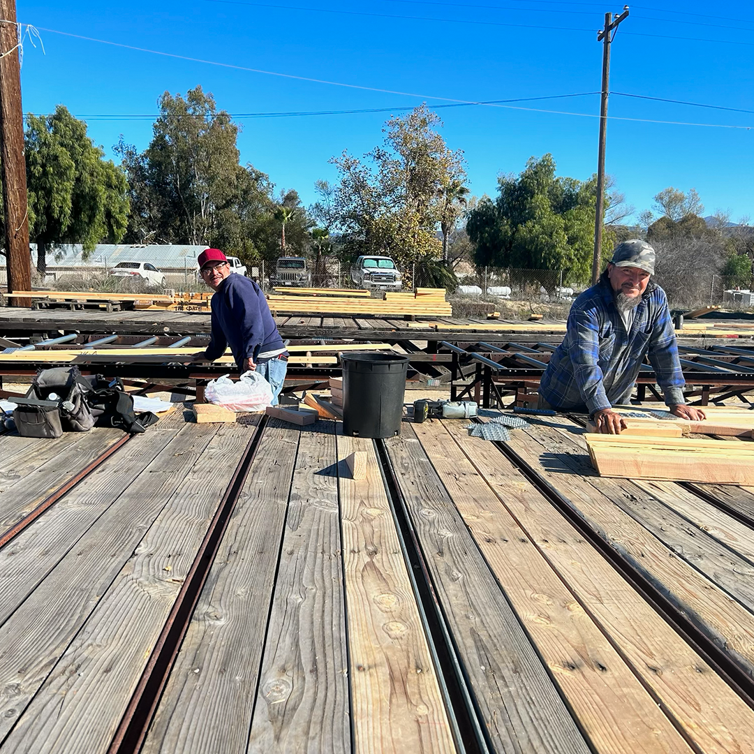 Two men working on wooden planks in an outdoor setting with train tracks. Blue sky.