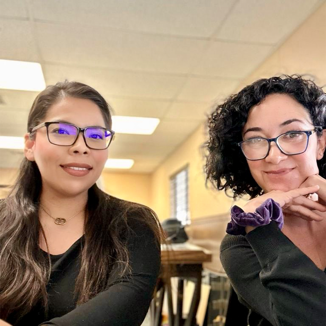 Two women smiling, wearing glasses, in a light-filled interior. One has dark hair, the other has curly hair.