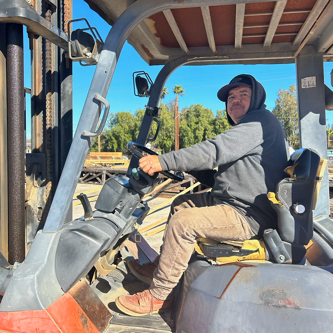 Man driving a forklift outdoors, wearing a hoodie and work pants.