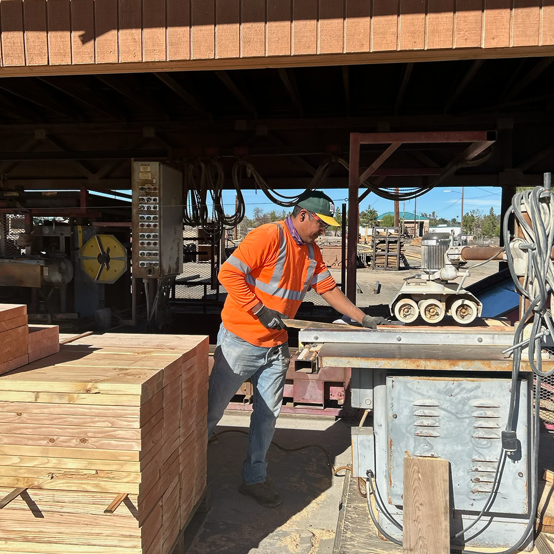 Man in orange safety vest working with machinery in a lumberyard, near stacks of wood.