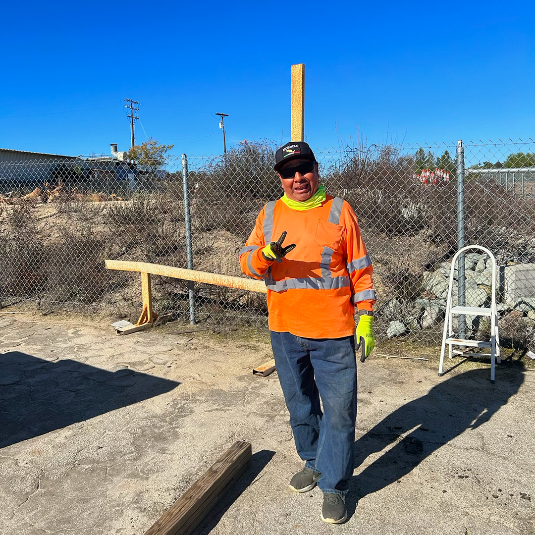 Man in orange safety gear standing outdoors with wood beams, making a peace sign.