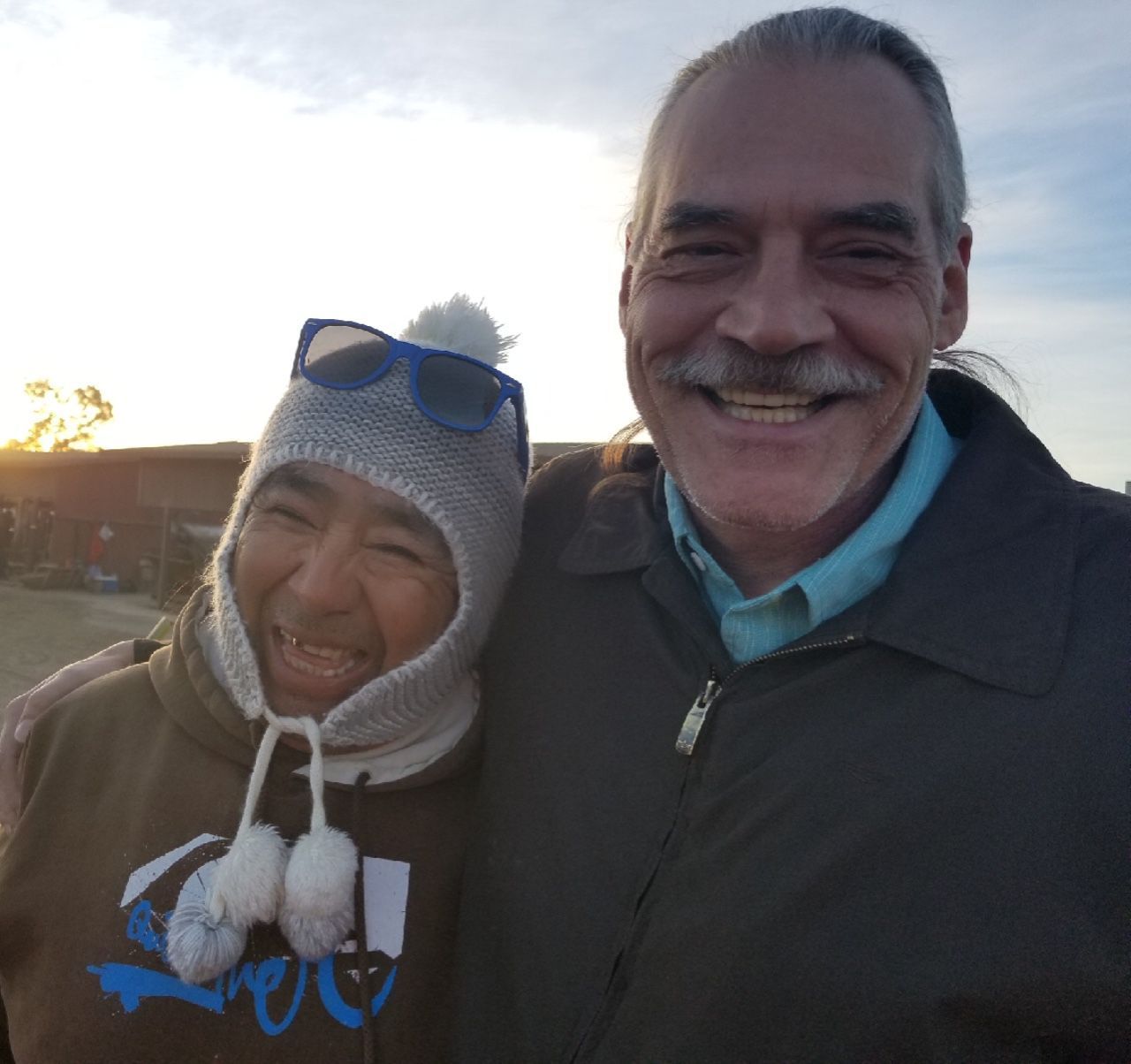 Two smiling men, one in a knit hat and sunglasses, the other with a mustache, standing outdoors.
