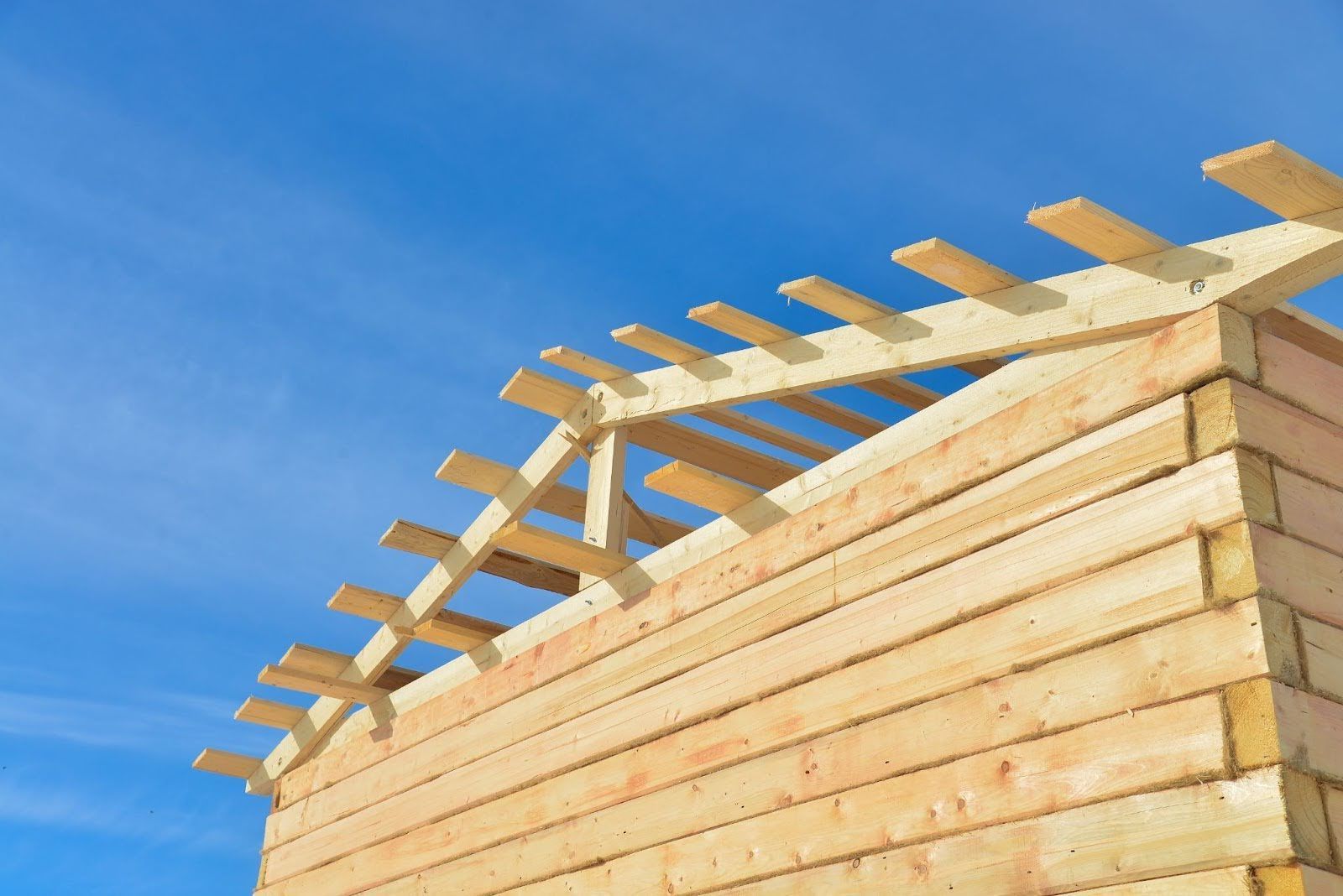 Wooden house frame under construction against a blue sky, roof rafters in place.