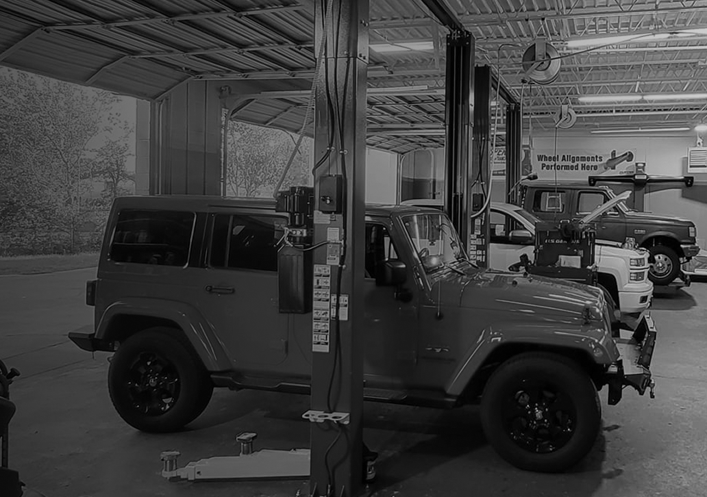 A black and white photo of a jeep on a lift in a garage. | Pearland Automotive