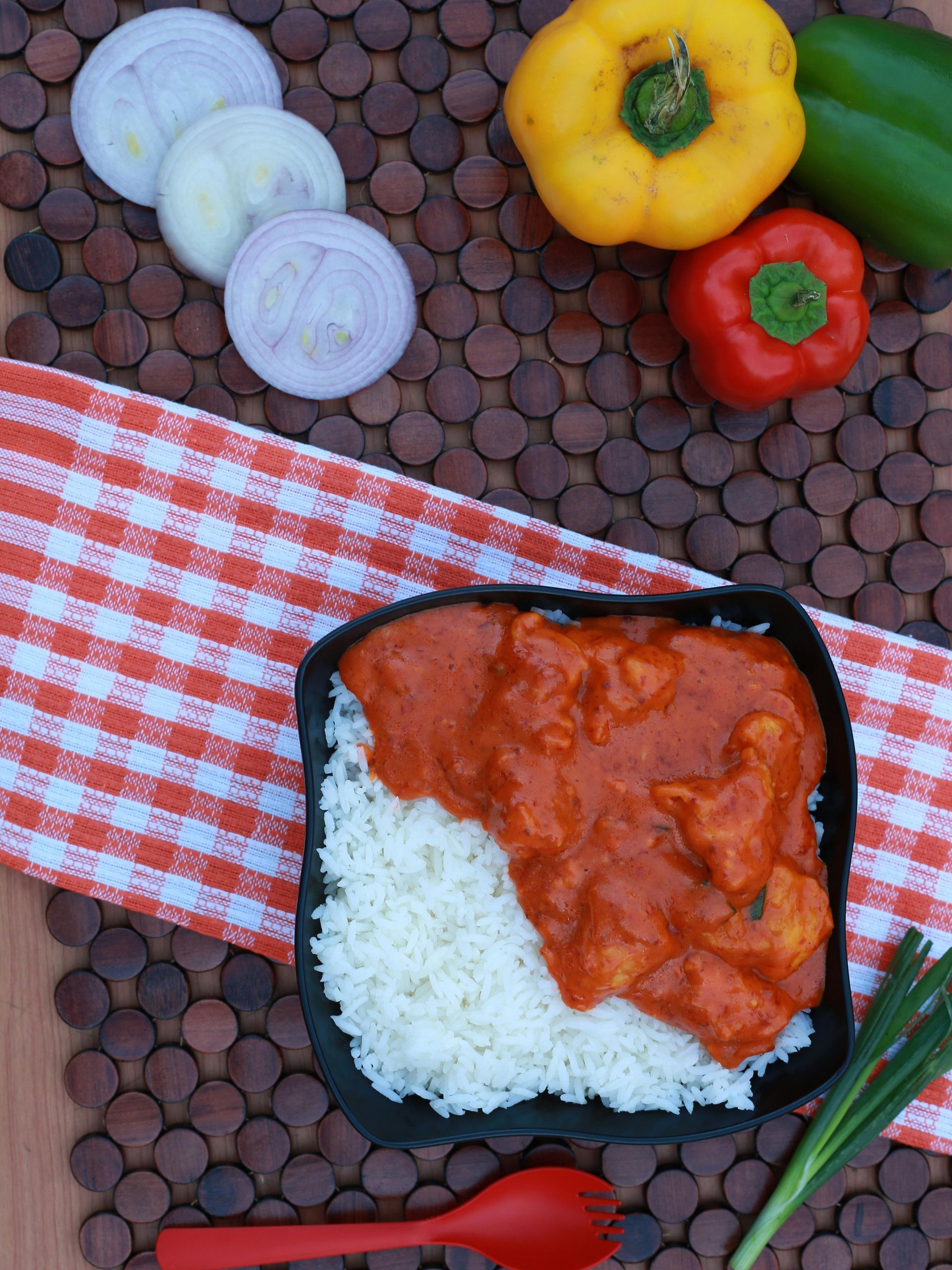 Chicken tikka masala with rice in a black bowl, red sauce, next to sliced onions, peppers and a red spoon.