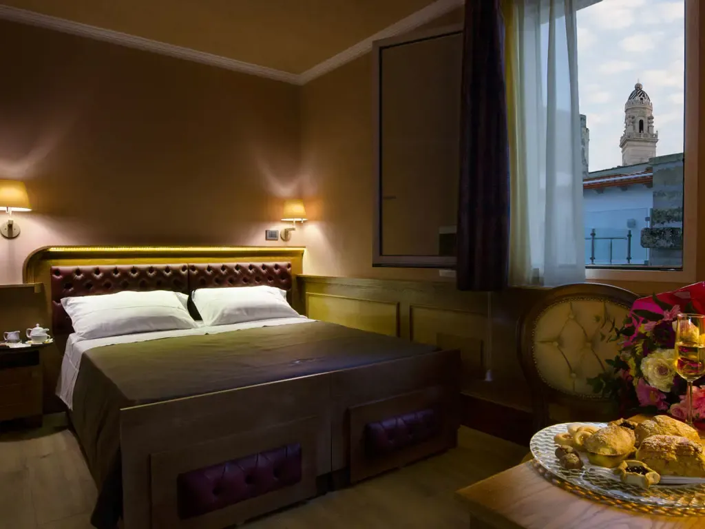 Bedroom with double bed, nightstands, window with view of a building, and table with flowers and pastries.
