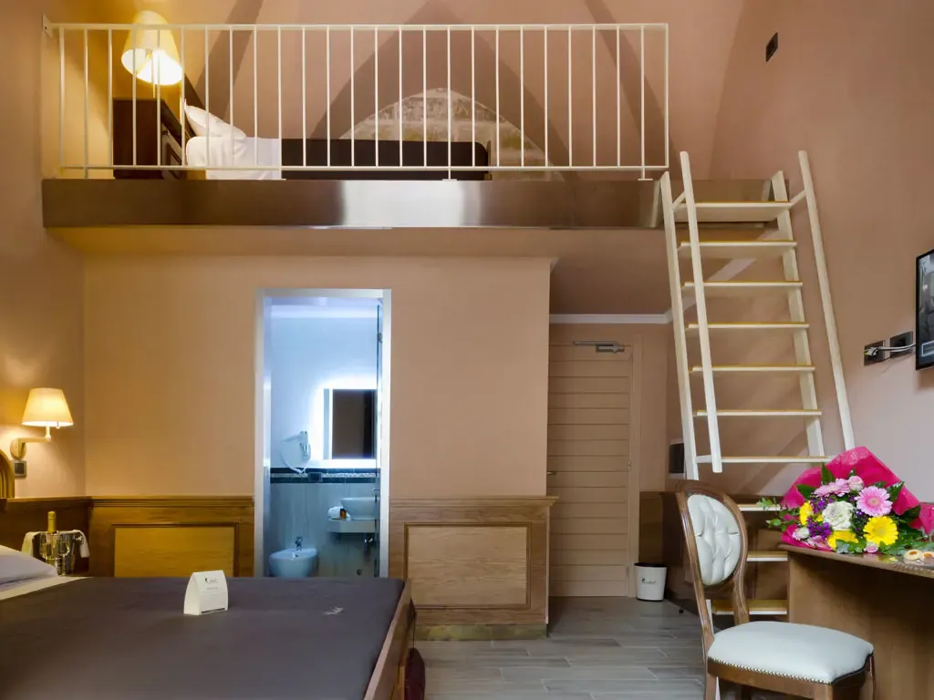 Hotel room with a loft bed, ladder, and bathroom visible. Pink walls, wooden furniture, and flowers on a desk.