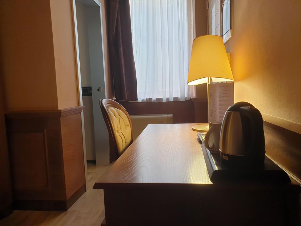 A hotel room desk with a lamp, a kettle, and a chair near a window with curtains.