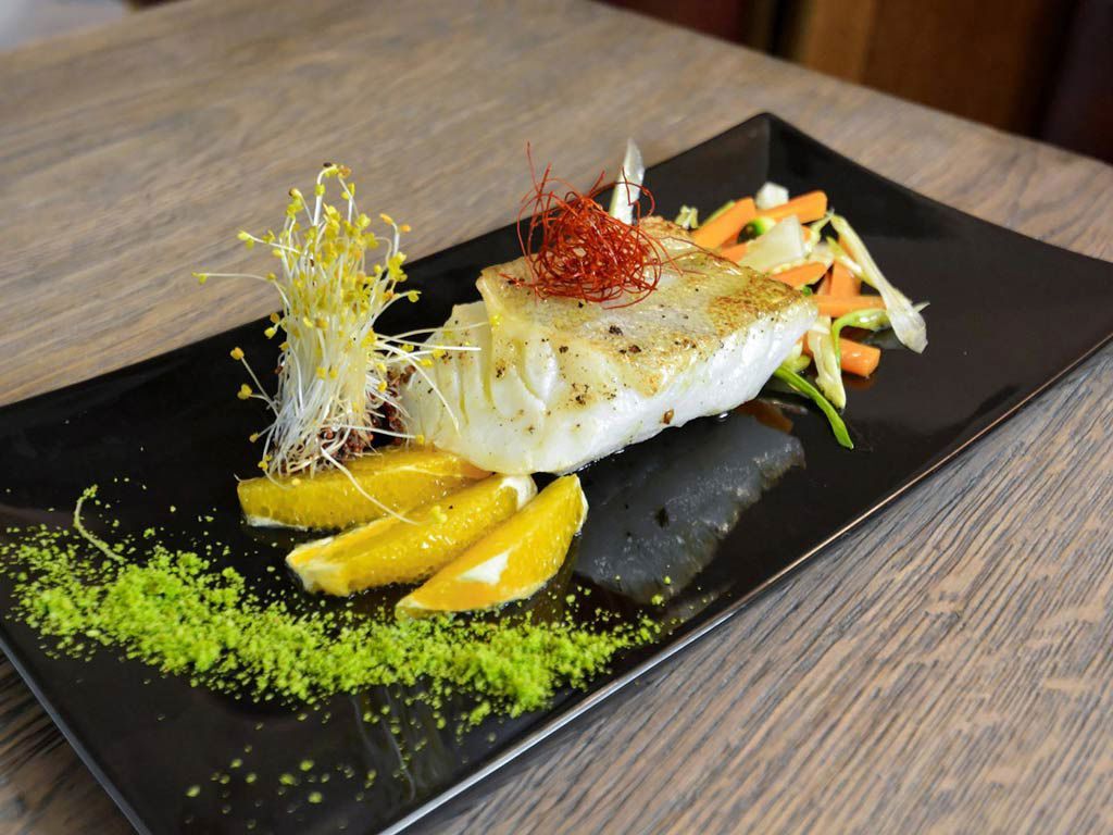 Grilled fish on a black plate with orange slices, vegetables, and green powder; served on a wood table.