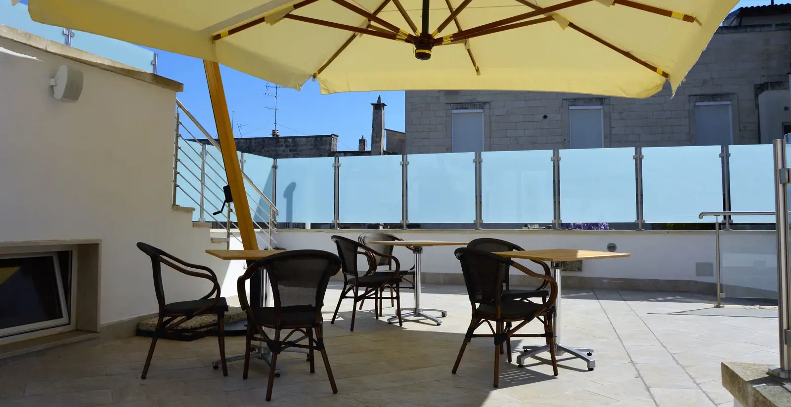 Rooftop patio with umbrella, tables, chairs, and frosted glass railing. Sunny day, city buildings in background.