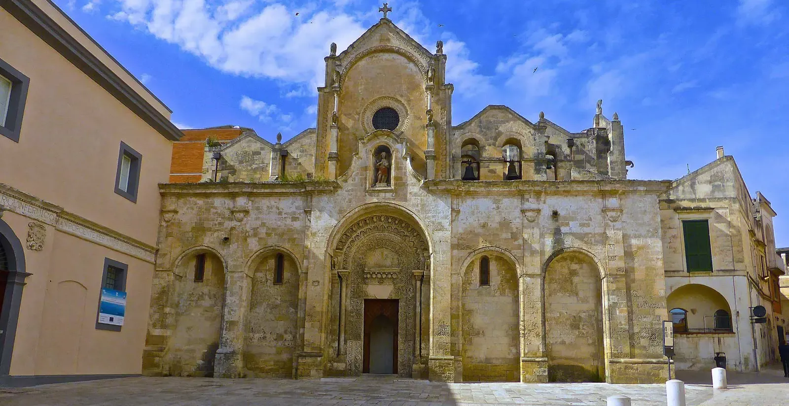 Una chiesa in pietra usurata dal tempo, con un ingresso ad arco e una croce sul tetto, sotto un cielo azzurro.