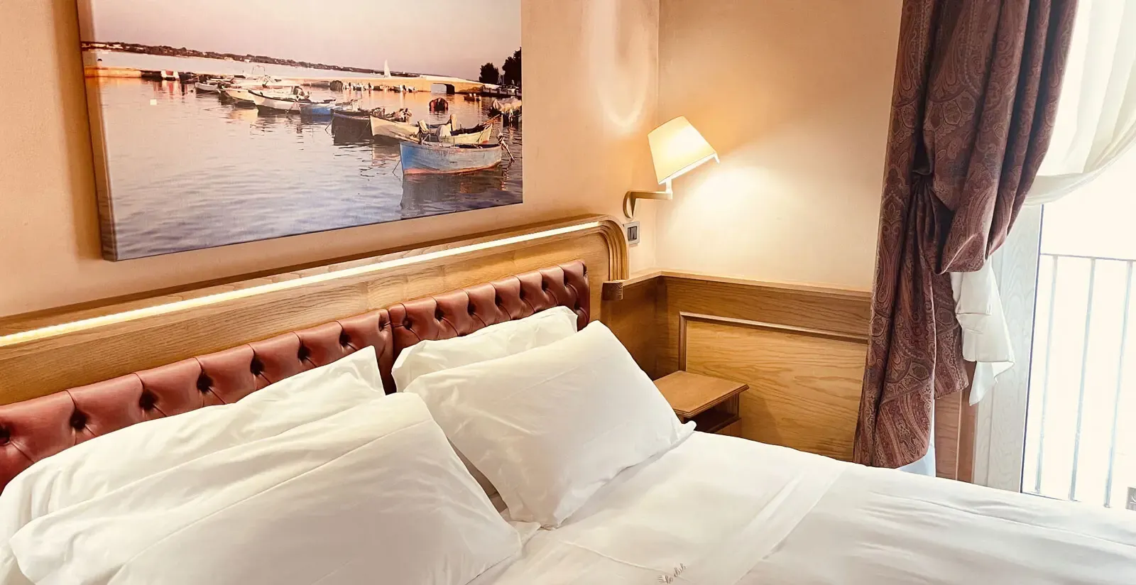 Bedroom with a double bed, white bedding, pink headboard, artwork above. Curtains and a balcony visible.