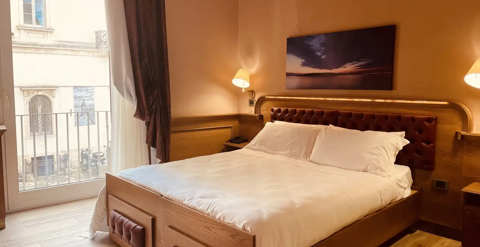 Hotel bedroom with a bed, artwork, and window overlooking a street.