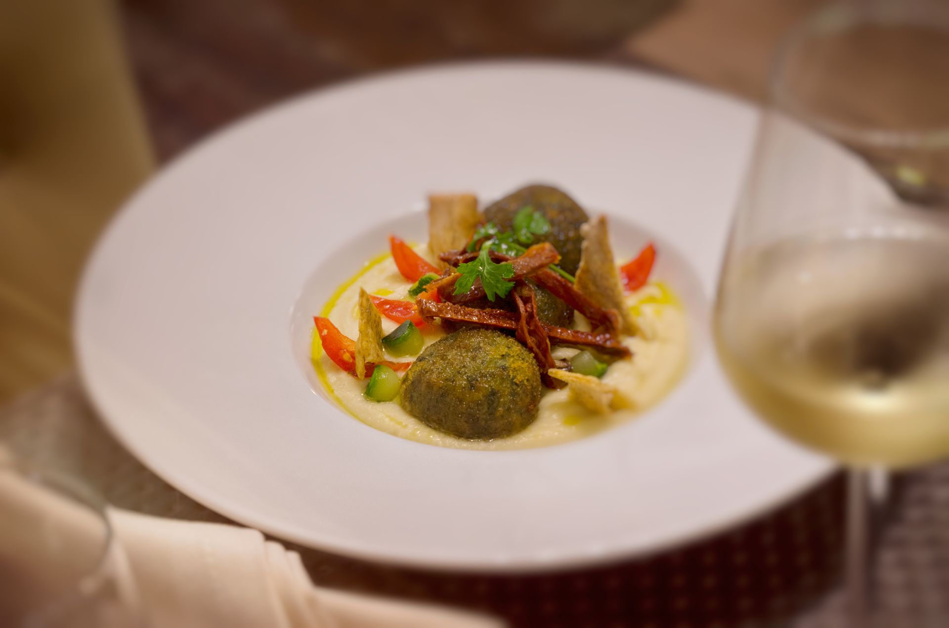 Falafel with garnishes on creamy base, white plate, and glass of white wine.