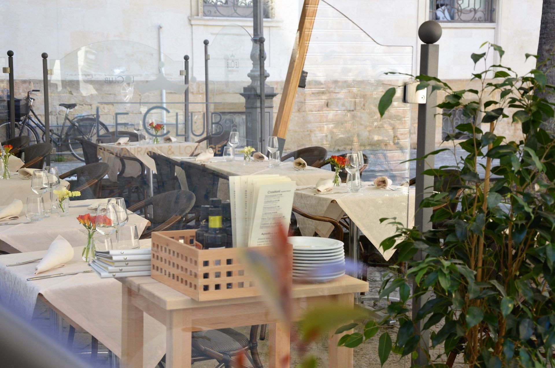 Outdoor restaurant tables set for dining, behind a glass window, street setting.