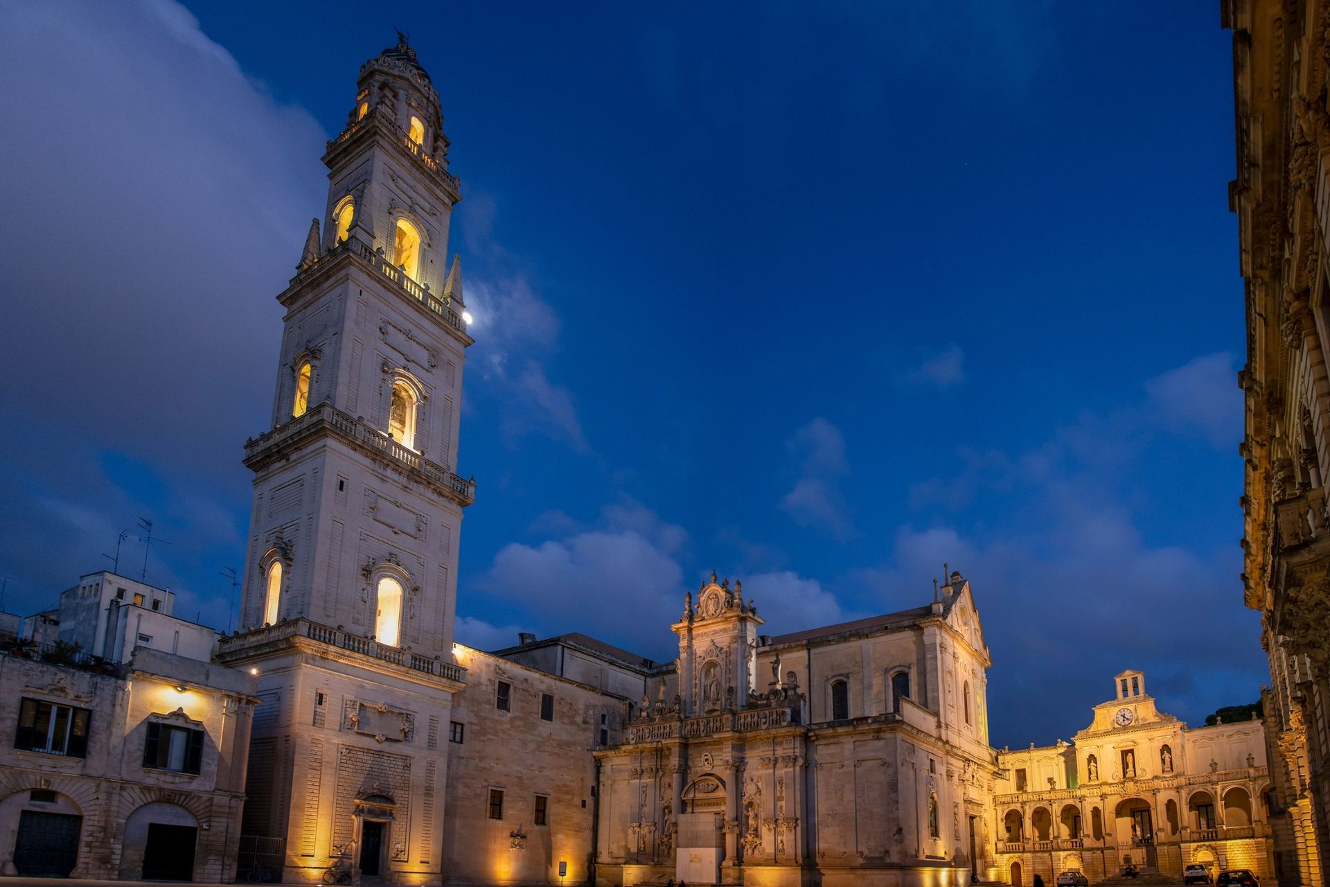 Vista notturna di un alto campanile illuminato e di un complesso edilizio a Lecce, in Italia, sotto un cielo blu intenso.