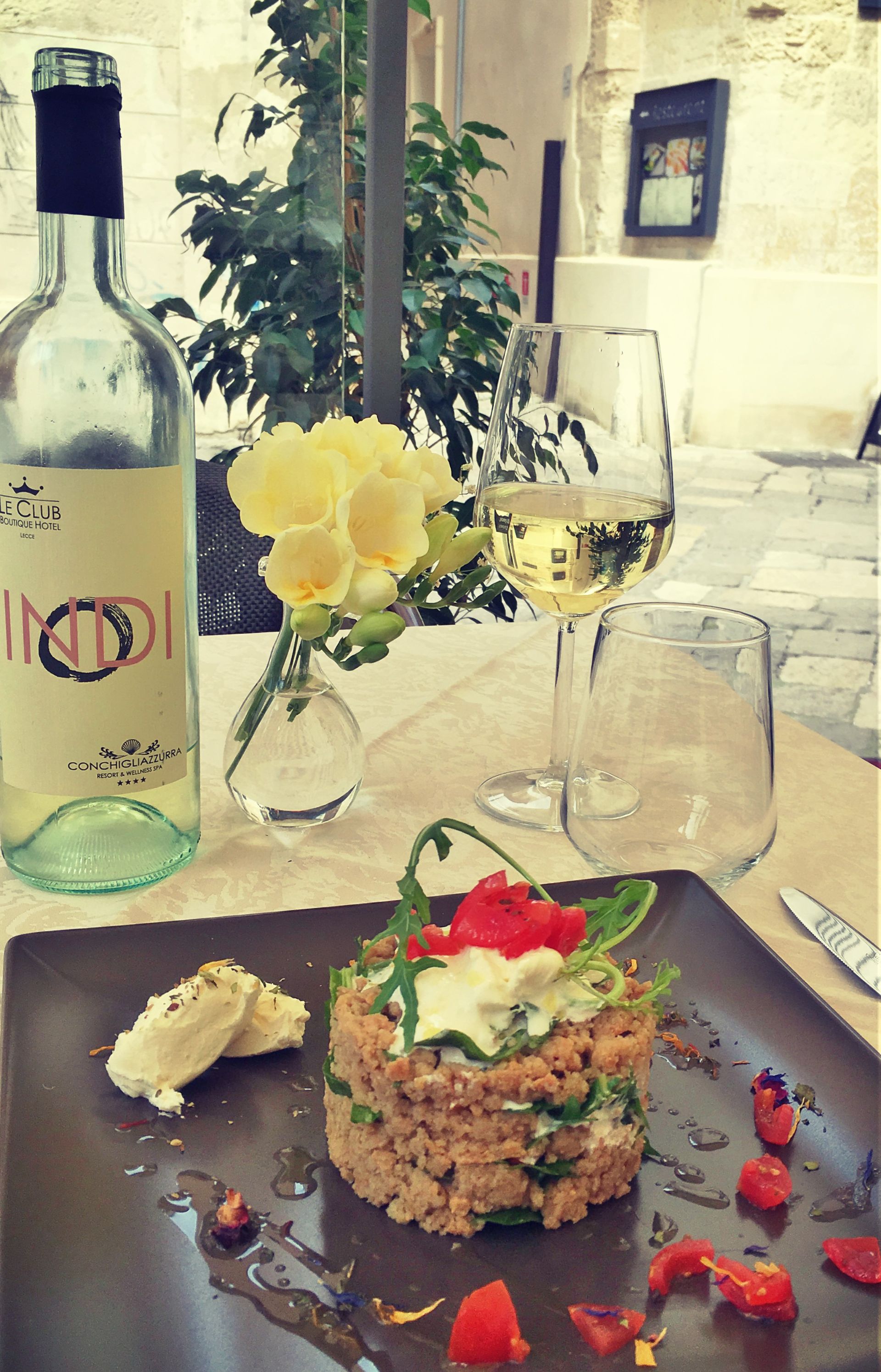 Tavolo apparecchiato con una bottiglia di vino, un bicchiere, cibo e fiori. All'aperto.