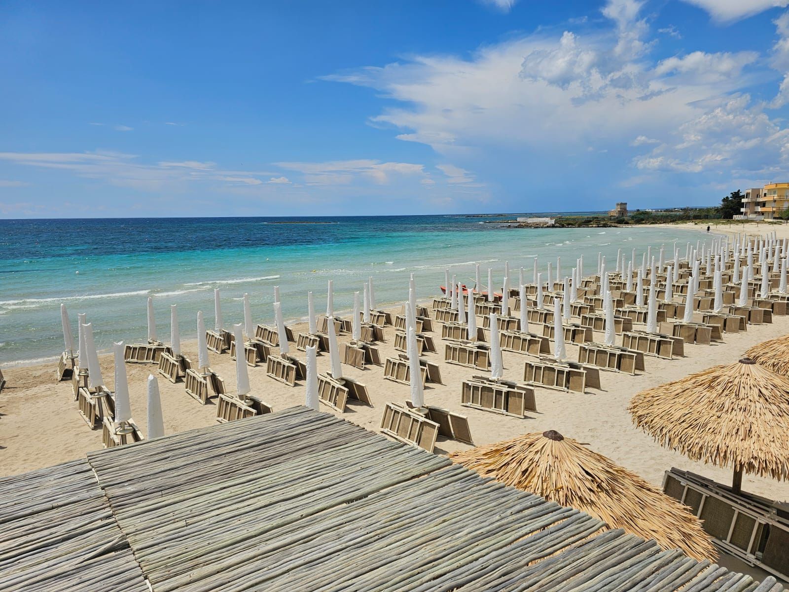 Beach scene: rows of chairs and white umbrellas on sandy shore, turquoise water, blue sky.