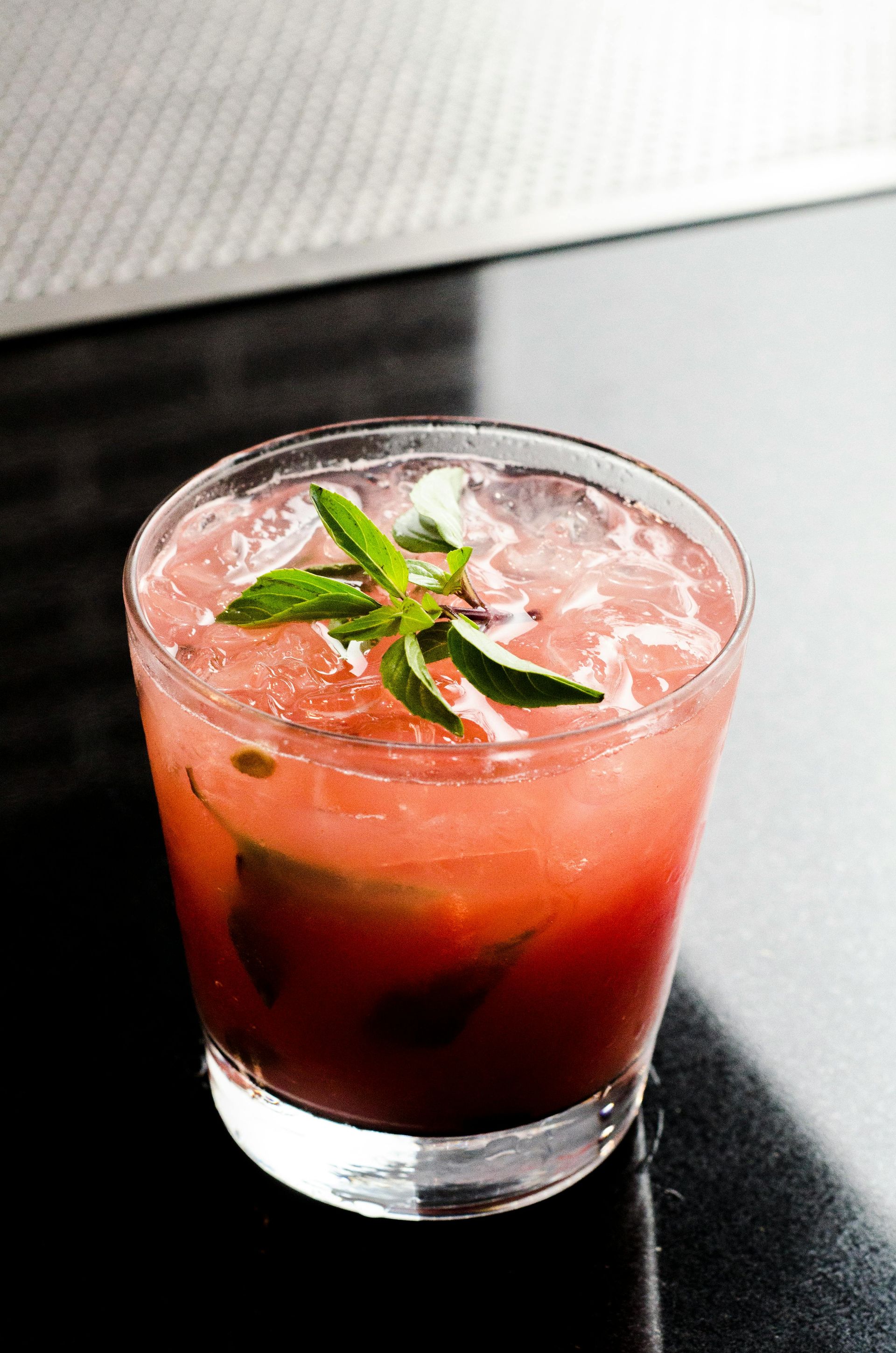 Pink cocktail in a glass with ice and mint garnish, on a dark surface.