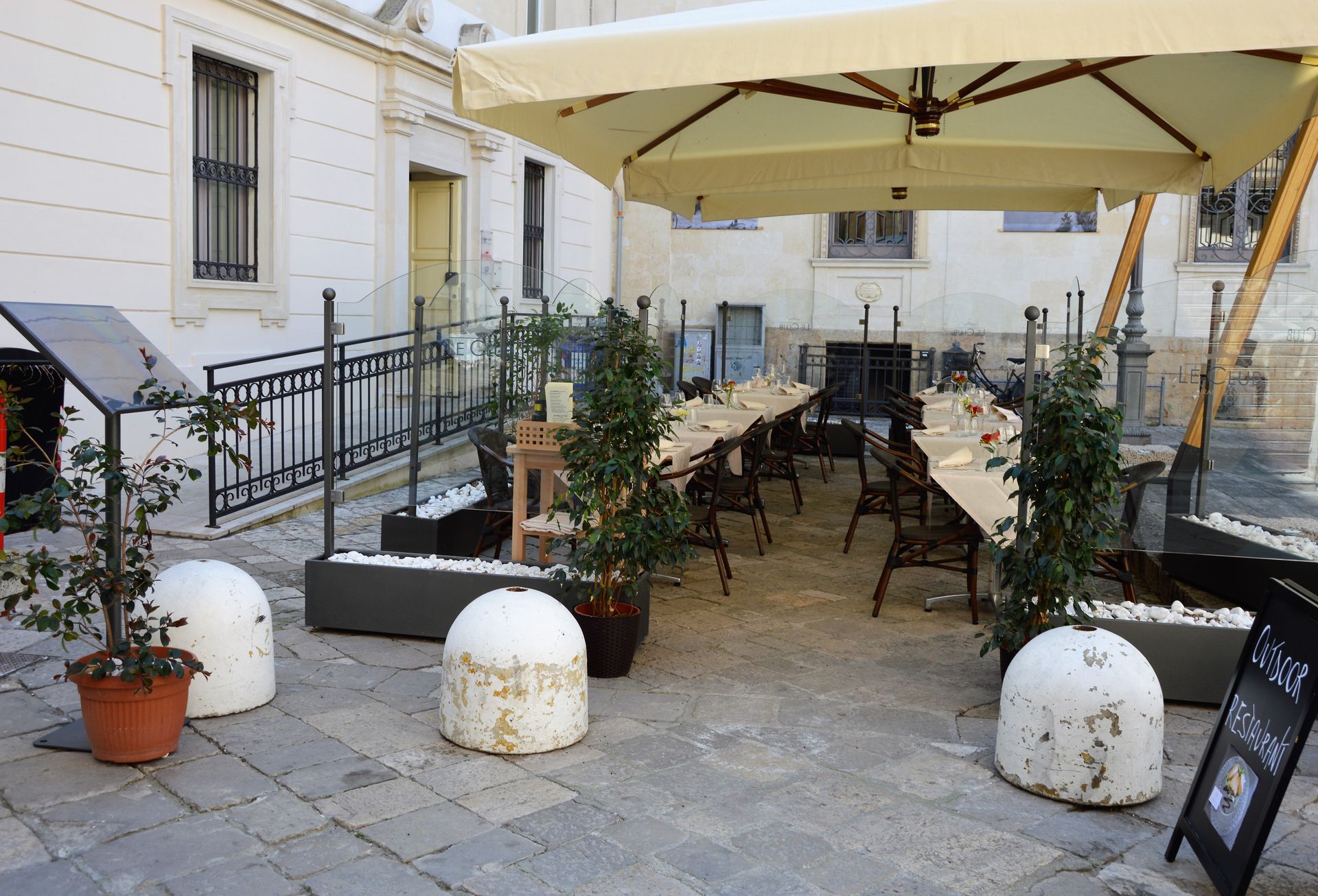 Outdoor restaurant seating area with tables set under umbrellas and trees. Buildings in the background.