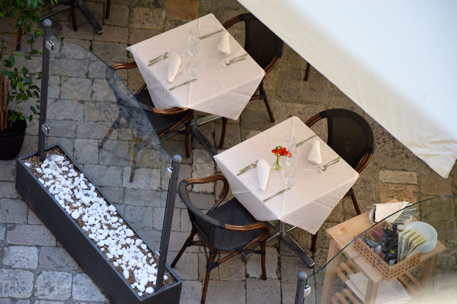 Overhead view of outdoor restaurant tables, set with white tablecloths, silverware, and napkins.