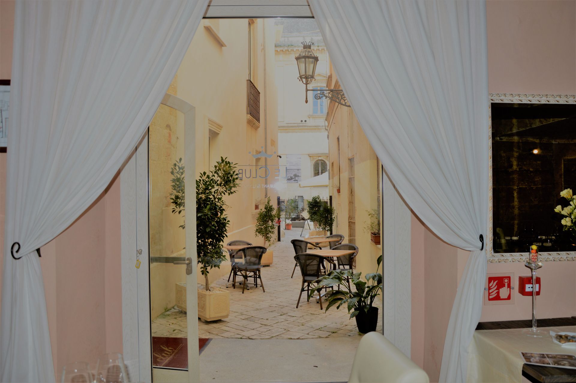 Open doorway revealing a courtyard with tables, chairs, and plants. Light-colored walls and buildings.