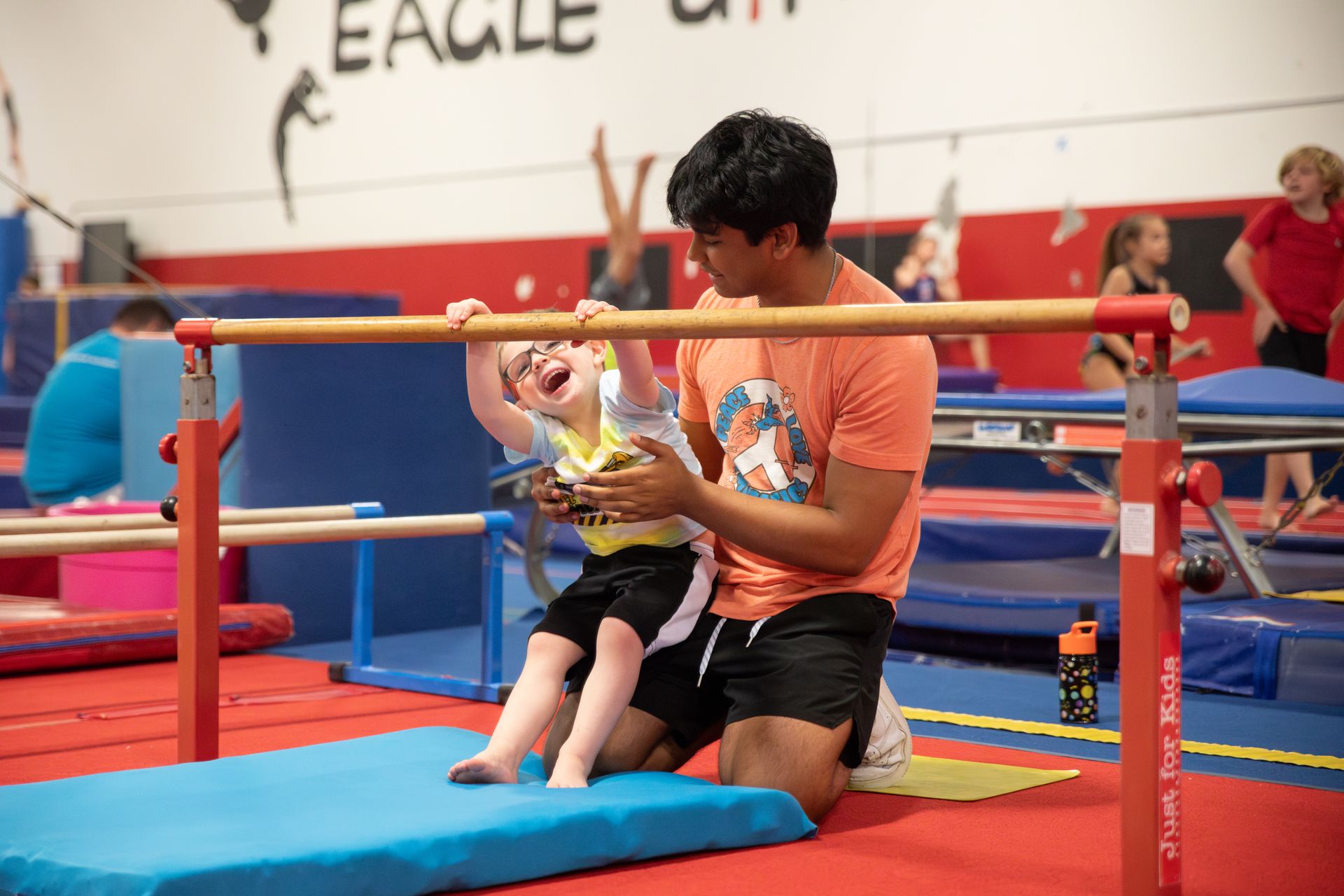 A man is kneeling down next to a child on a parallel bars in a gym.