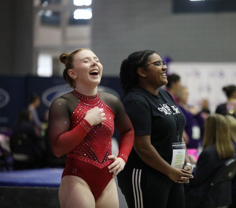 Two female gymnasts are laughing together while standing next to each other in a gym.