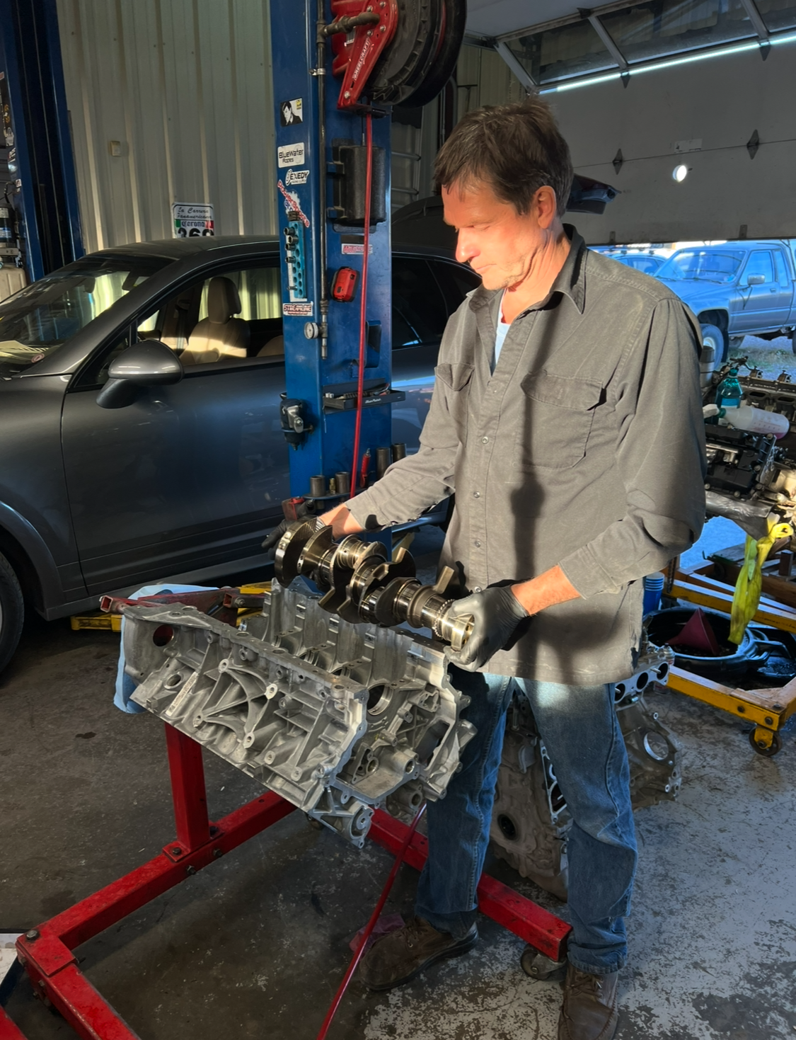 Mechanic in a garage holding engine parts, examining them. Gray shirt, blue jeans, standing near a car.