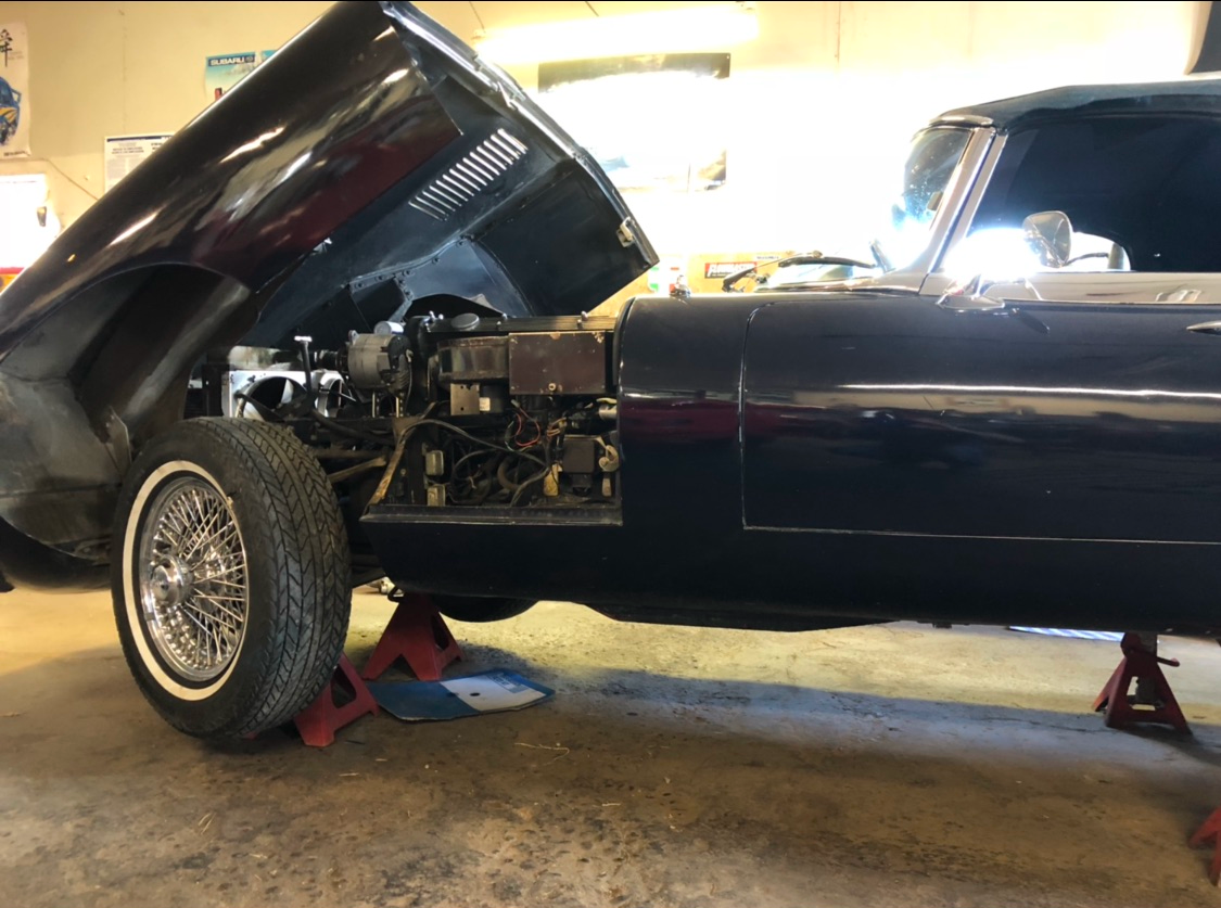 Black Jaguar E-type with hood open, raised on jack stands in a garage, showing the engine and front wheel.