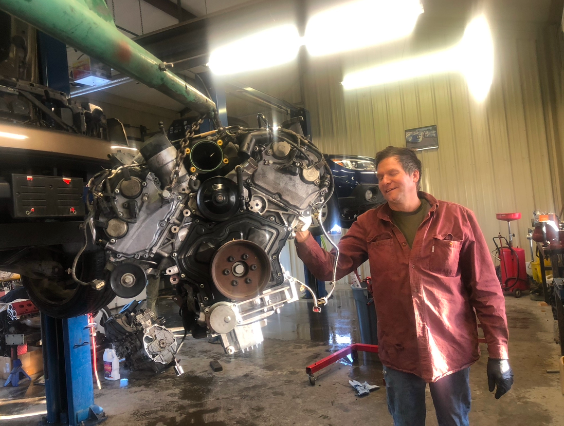 Man in a garage holding a large car engine. The engine is suspended, and the man smiles.