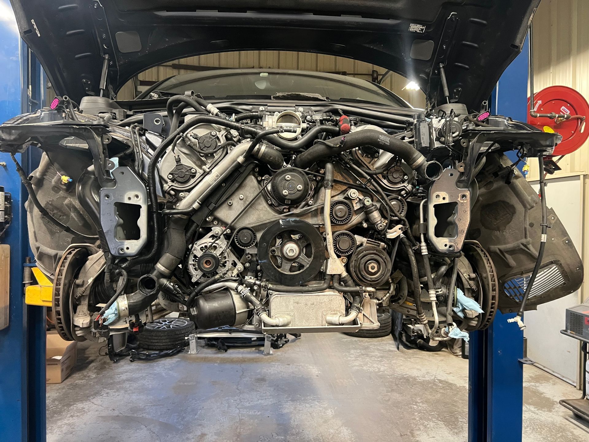 Car on a lift with hood open, revealing a complex engine and removed front end components in a repair shop.