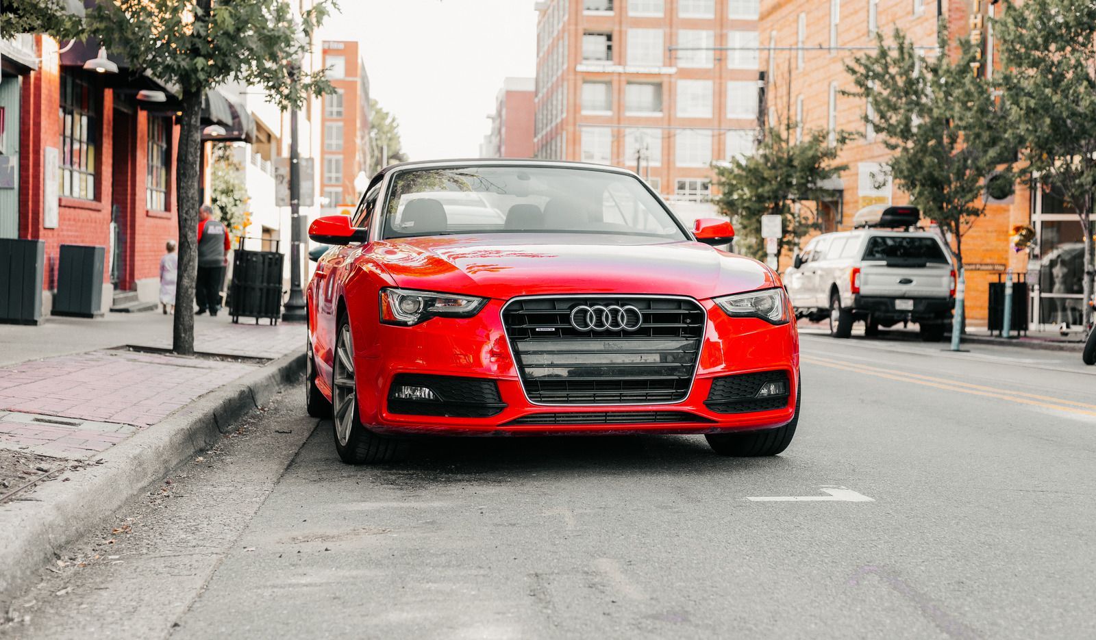 Red Audi car parked on a city street. Buildings, trees, and a white truck in the background.