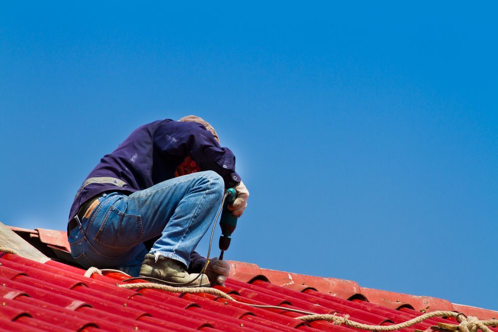 A Man is Kneeling on a Roof Using a Drill — Roof Restoration Masters In Rutherford, NSW