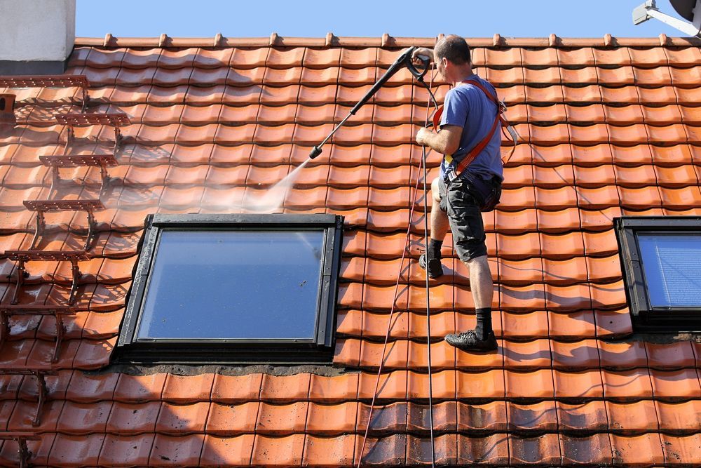 A Man is Cleaning the Roof of a House With a High Pressure Washer — Roof Restoration Masters In Boolaroo, NSW