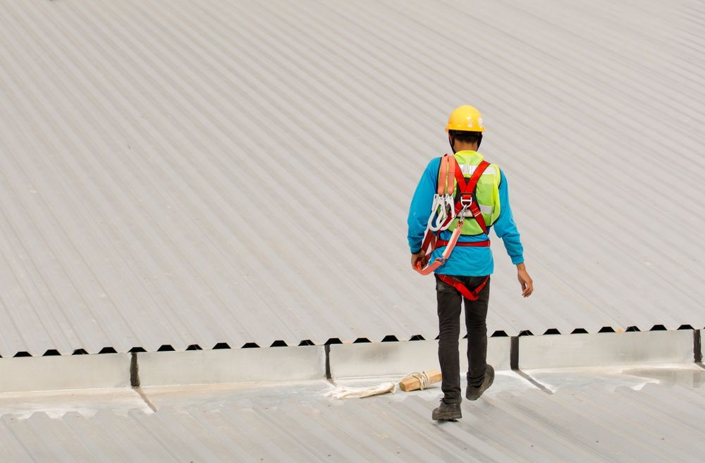 A Man Wearing a Hard Hat and Safety Harness is Walking on a Roof — Roof Restoration Masters In Raymond Terrace, NSW