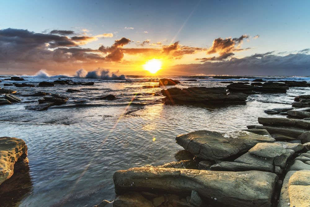 The Sun is Setting Over the Ocean With Rocks in the Foreground — Roof Restoration Masters In Bateau Bay, NSW
