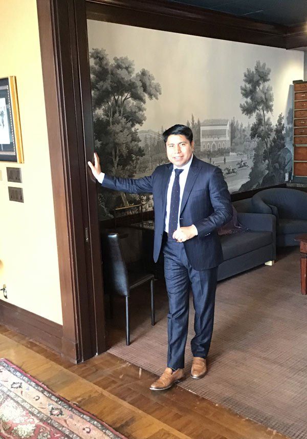Man in navy suit, brown shoes, poses in a room with mural and dark wood trim.