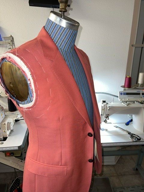 A coral-colored blazer on a tailor's mannequin, mid-construction, with a sewing machine in the background.