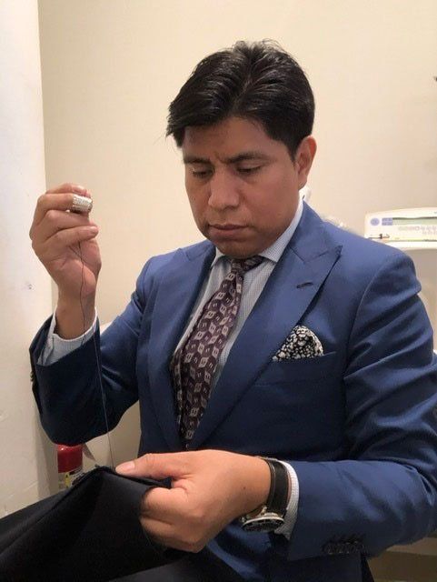 Man in a blue suit sewing black fabric. He is holding a needle and focused.