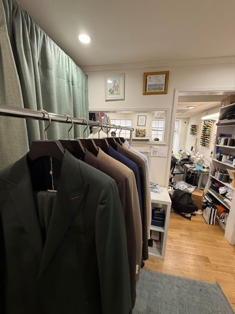 Suits hanging on a rack in a tailoring shop. Light green curtains, visible doorway to another room.