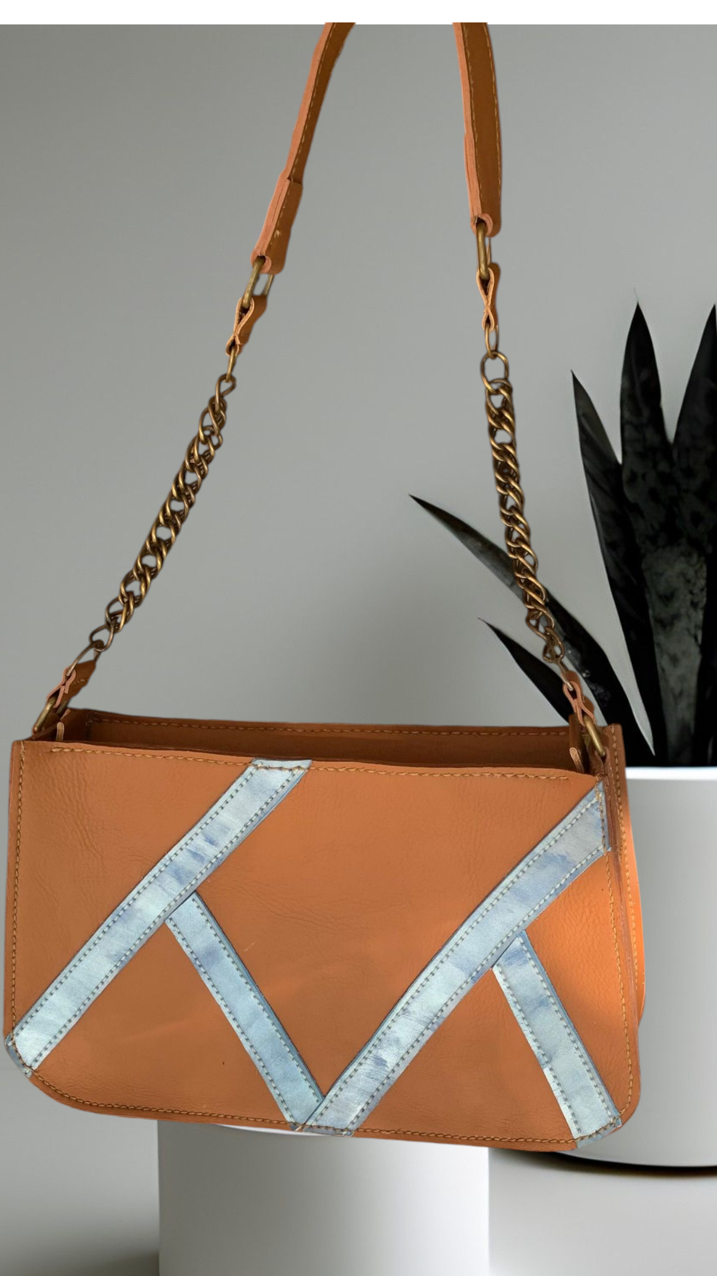 Bolso marrón y plateado con asa de cadena, expuesto delante de una planta.