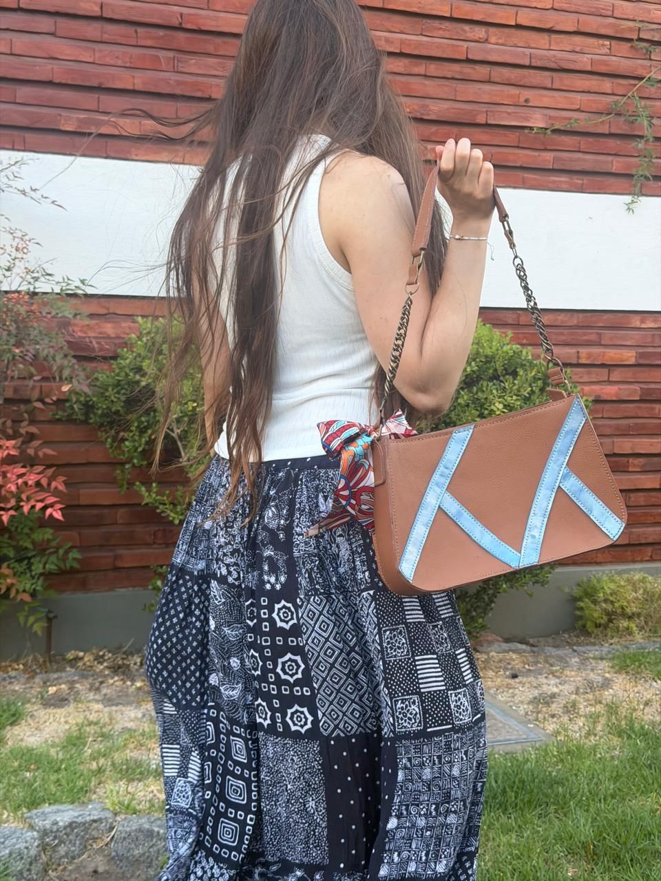 Bolso marrón y plateado con asa de cadena, expuesto delante de una planta.