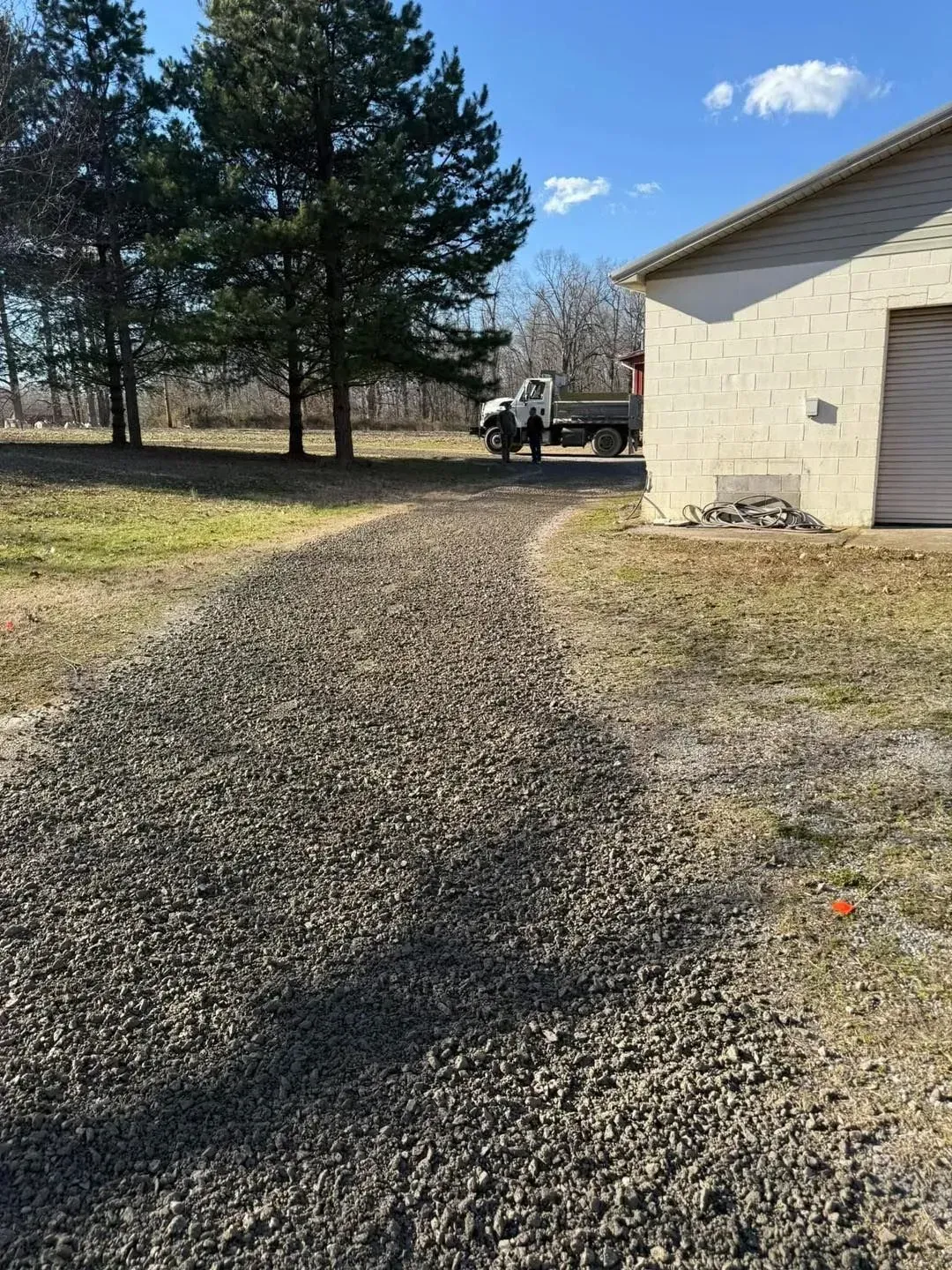 Gravel driveway beside a beige house, with trees and a small utility vehicle in the distance under a blue sky.