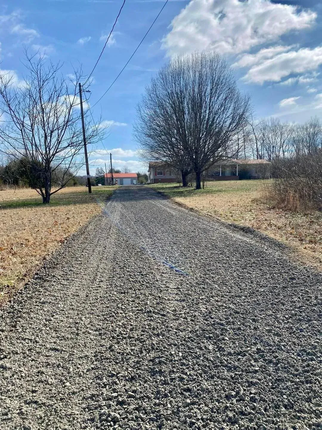 Gravel driveway leading to a rural house, lined with bare trees under a partly cloudy blue sky.