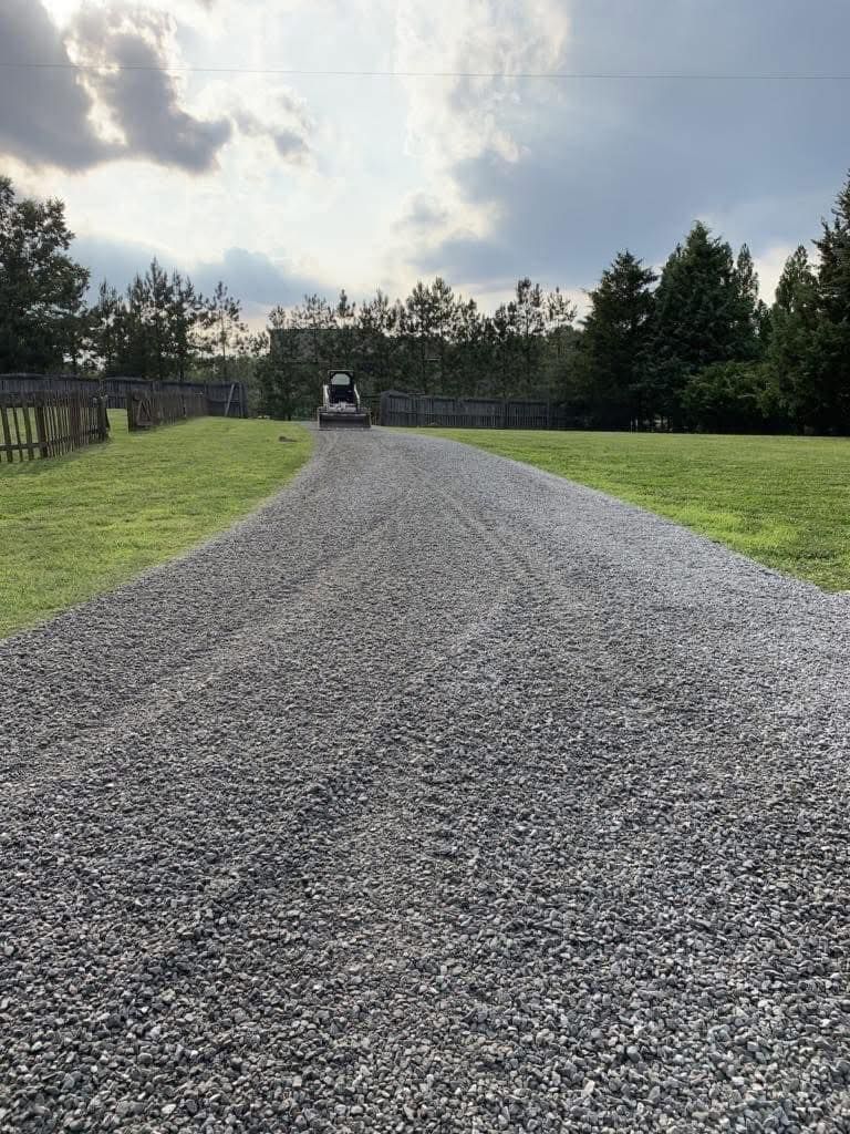 Gravel driveway leading to a distant golf cart under a cloudy sky, bordered by trees and grass.